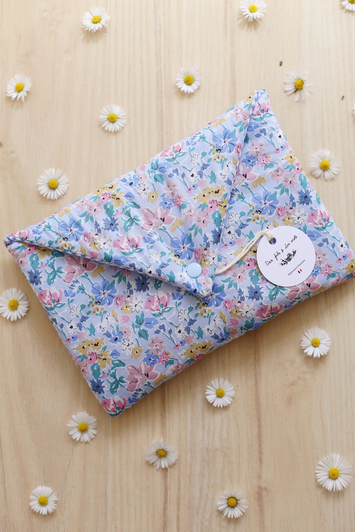 Housse pour livre en Liberty of London imprimé fleurs bleues et roses pastel, fermée avec bouton pression bleu, étiquette Des fils et des mots, entourée de marguerites sur parquet bois
