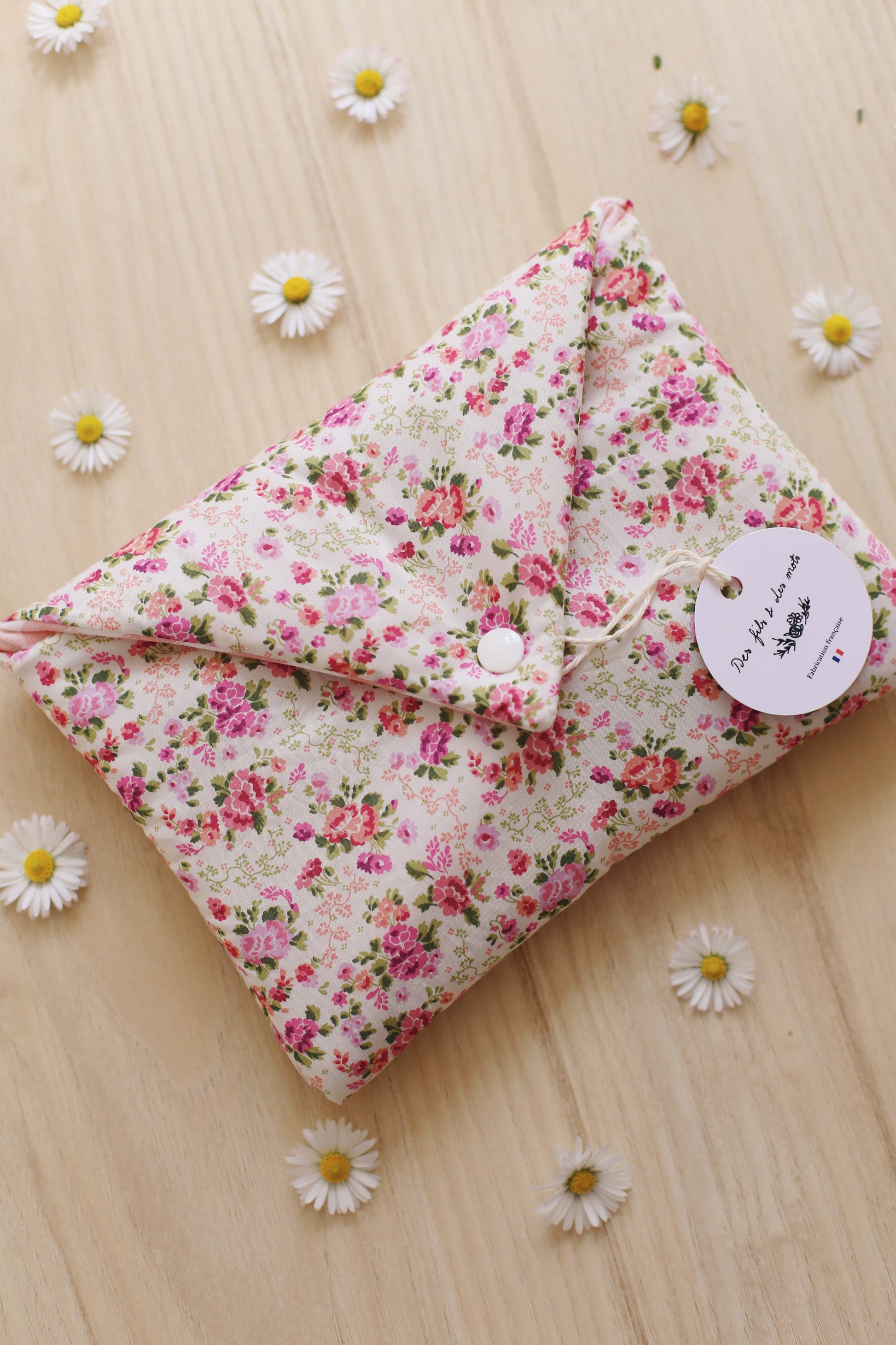 Housse pour livre artisanale en Liberty of London imprimé roses roses sur fond crème, fermée avec bouton pression blanc, étiquette ronde Des fils et des mots, entourée de marguerites sur parquet bois