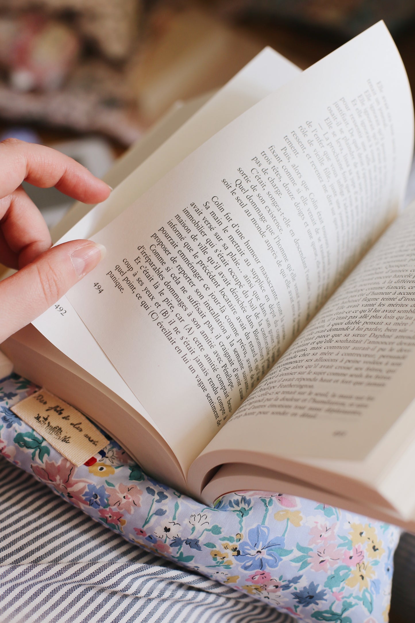 Livre ouvert en français tenu à la main, posé sur La Doucette coussin de lecture en Liberty of London fleuri bleu et jaune, utilisé sur un canapé rayé, étiquette Des fils et des mots visible