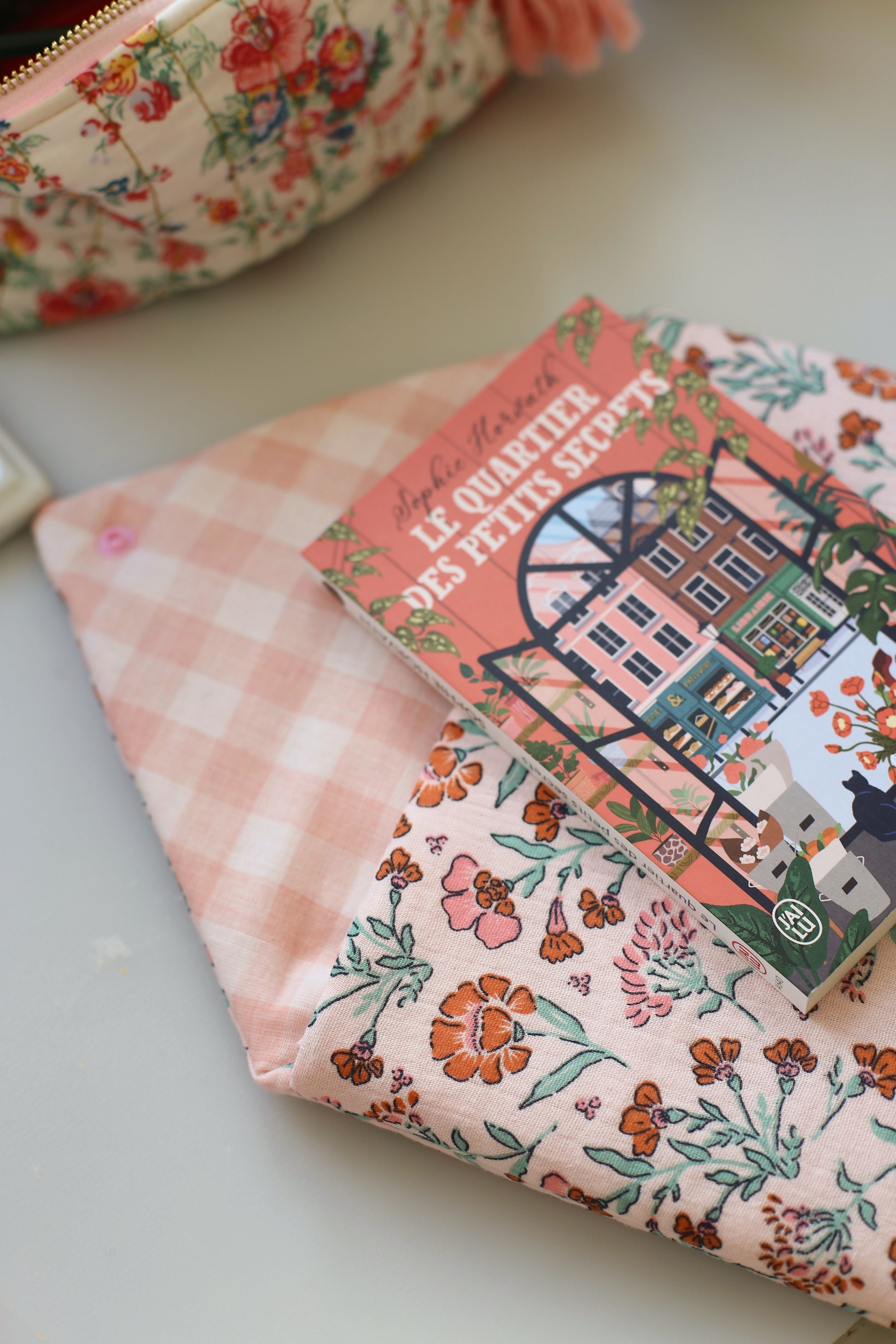 Housse pour livre de poche ouverte en double gaze rose à fleurs orangées, avec le roman Le Quartier des petits secrets de Sophie Horvath glissé à l'intérieur, doublure vichy rose visible