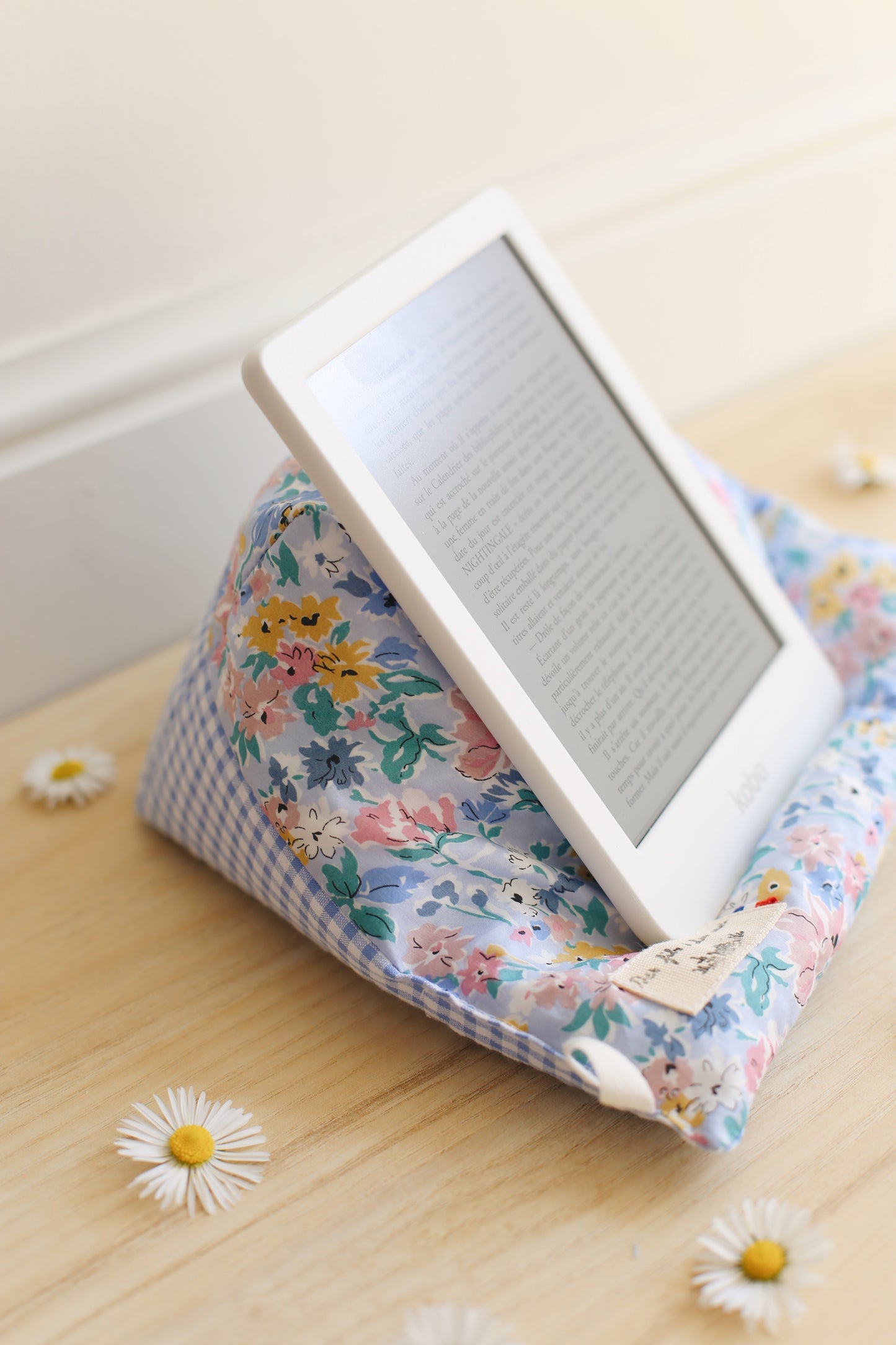 La Doucette coussin de lecture en Liberty of London fleuri bleu, jaune et rose, supportant une liseuse Kindle, posée contre un mur blanc sur parquet bois, marguerite au sol