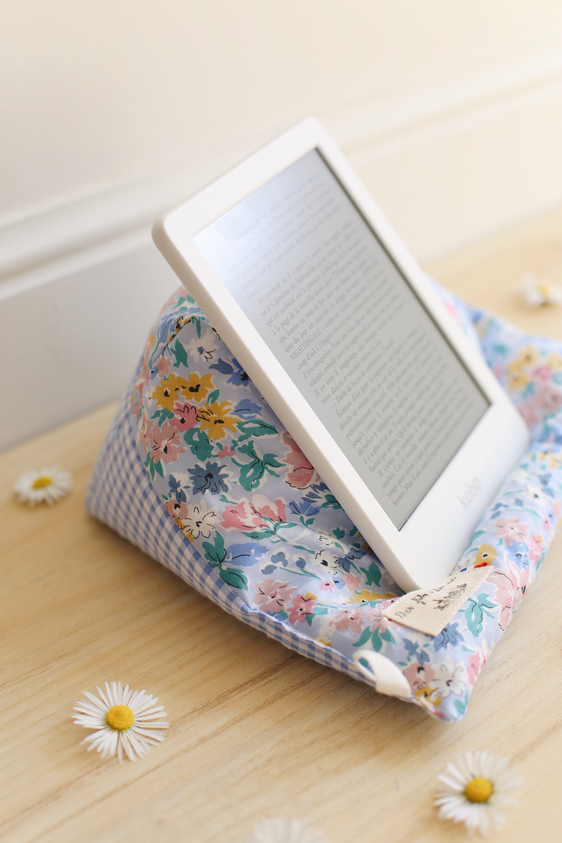 La Doucette coussin de lecture en Liberty of London fleuri bleu, jaune et rose, supportant une liseuse Kindle, posée contre un mur blanc sur parquet bois, marguerite au sol