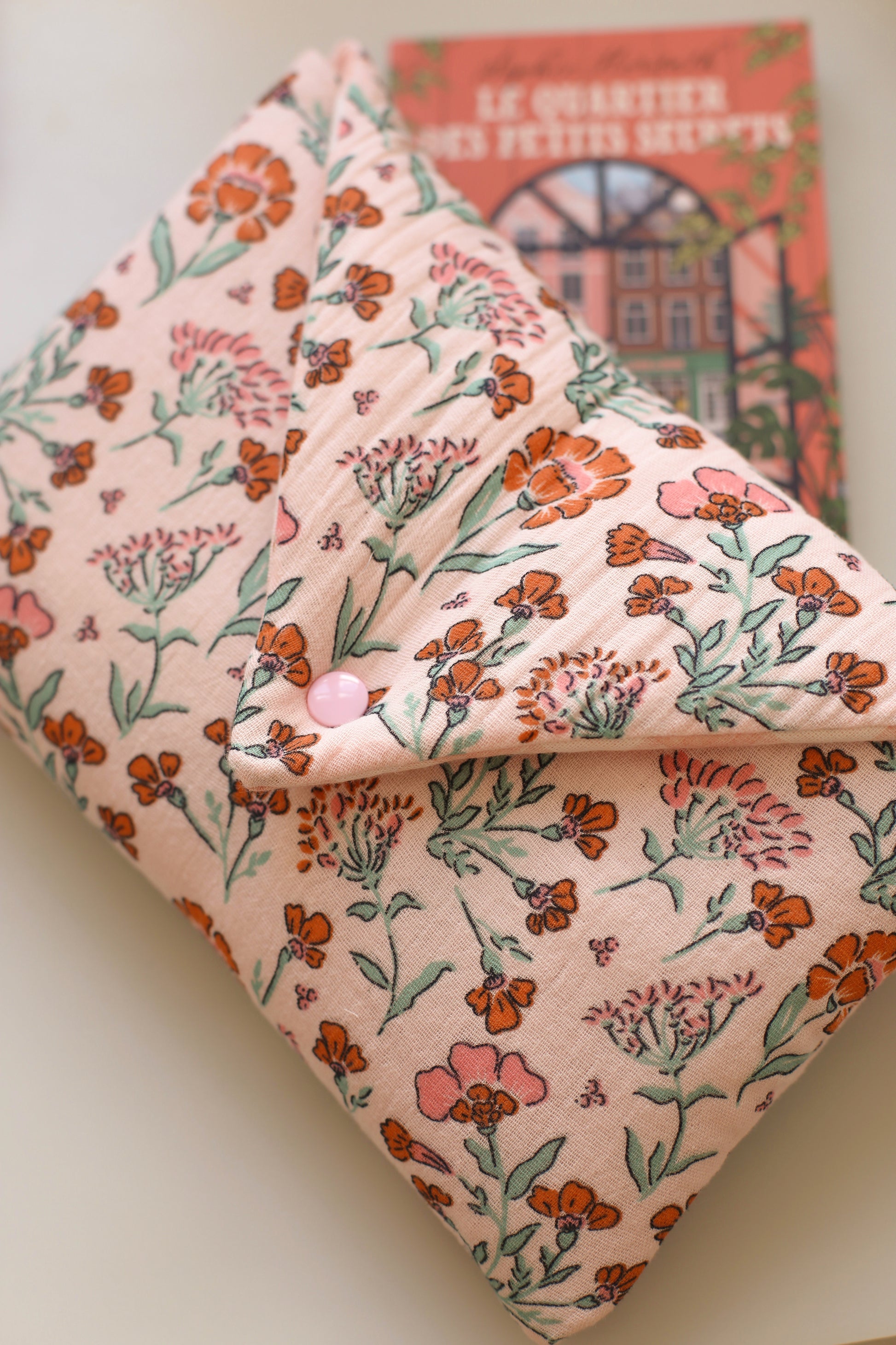 Housse pour livre en double gaze de coton rose à fleurs orangées et roses, fermée par un bouton pression, posée sur le roman Le Quartier des petits secrets de Sophie Horvath