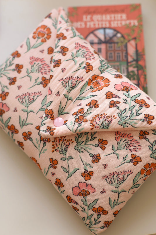 Housse pour livre en double gaze de coton rose à fleurs orangées et roses, fermée par un bouton pression, posée sur le roman Le Quartier des petits secrets de Sophie Horvath