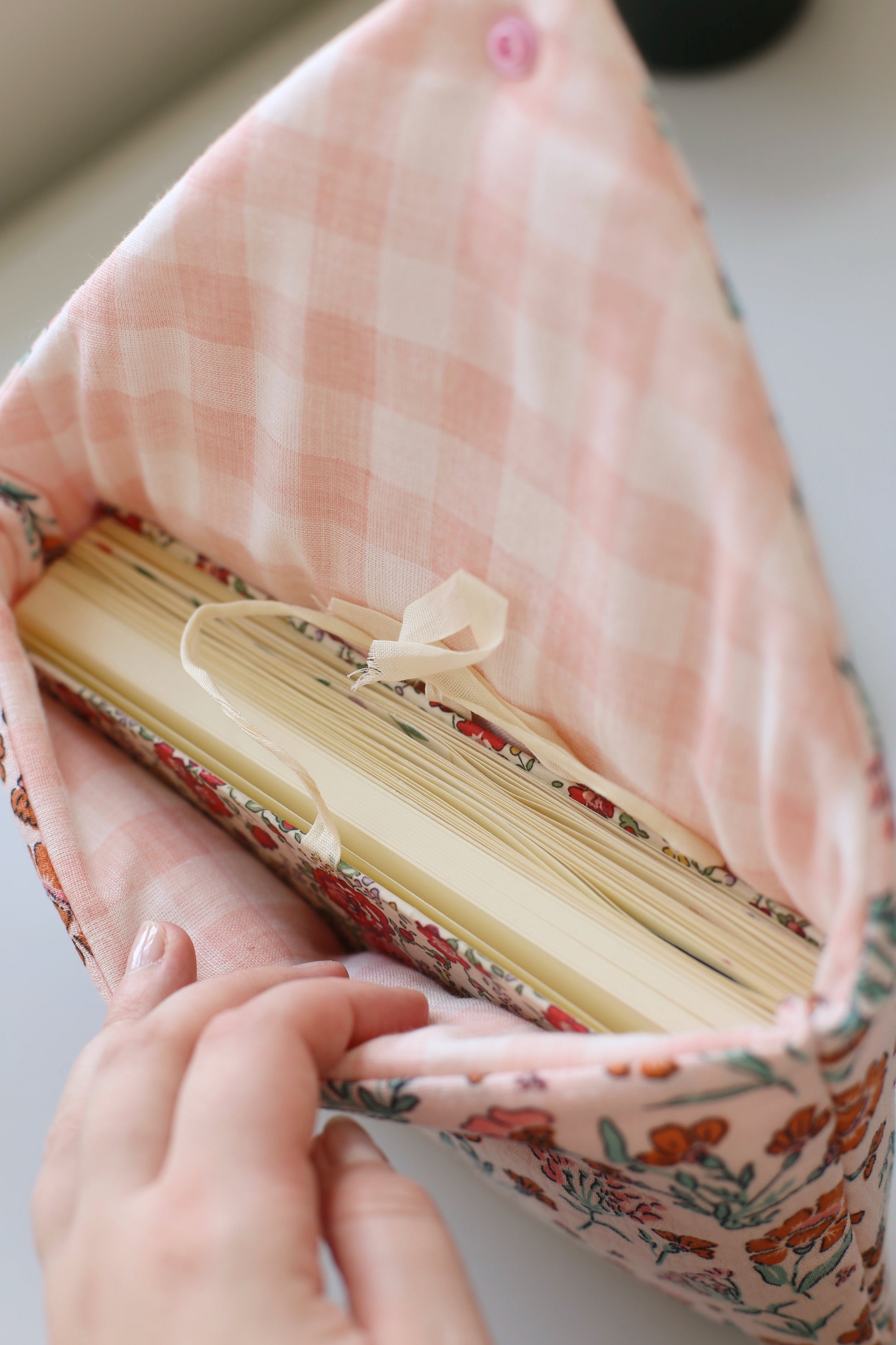 Intérieur de la housse pour livre ouverte, révélant la doublure en double gaze vichy rose et un livre avec un marque-page ruban, tenue dans les mains